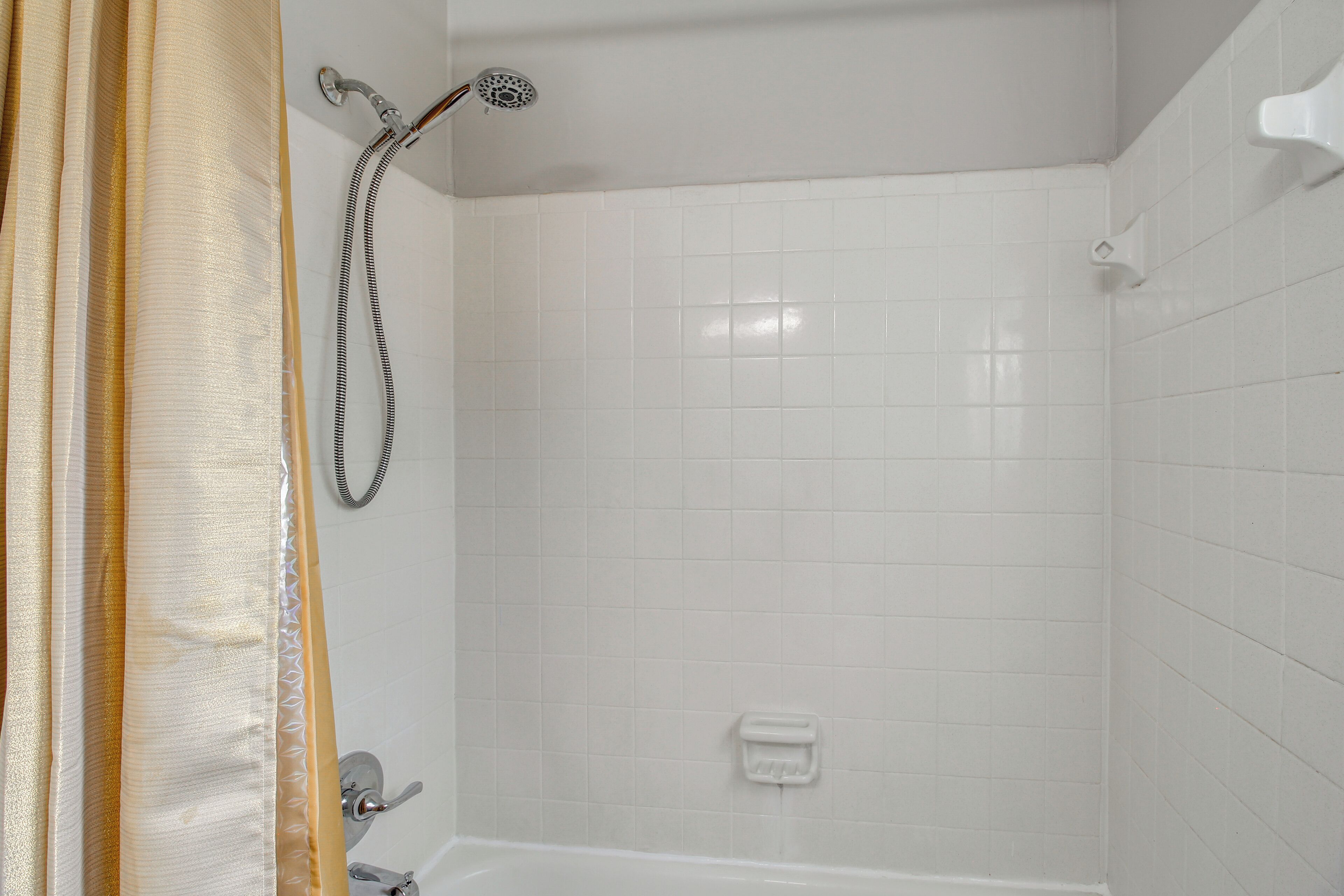 Ein Badezimmer mit weiß gefliesten Wänden, einer Dusche inklusive Handbrause und einer Badewanne.