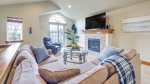 A spacious living room with a large sectional sofa, a glass coffee table, and a flat-screen TV mounted above a fireplace. - Walk to Lincoln City Beach: Townhome w/ Balconies! (Lincoln City)