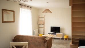 Ein Wohnzimmer mit einer Couch, einem Fernseher, einem kleinen Tisch, einer Leiter und einem Fenster mit Vorhängen.