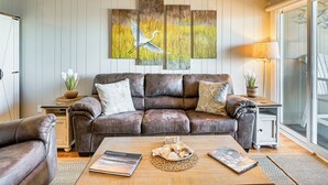A living room with a brown leather sofa, a wooden coffee table with decorative items, and a large wall painting of a bird in flight. - Waterfront Gem w/ Balcony in Garden City (Murrells Inlet)
