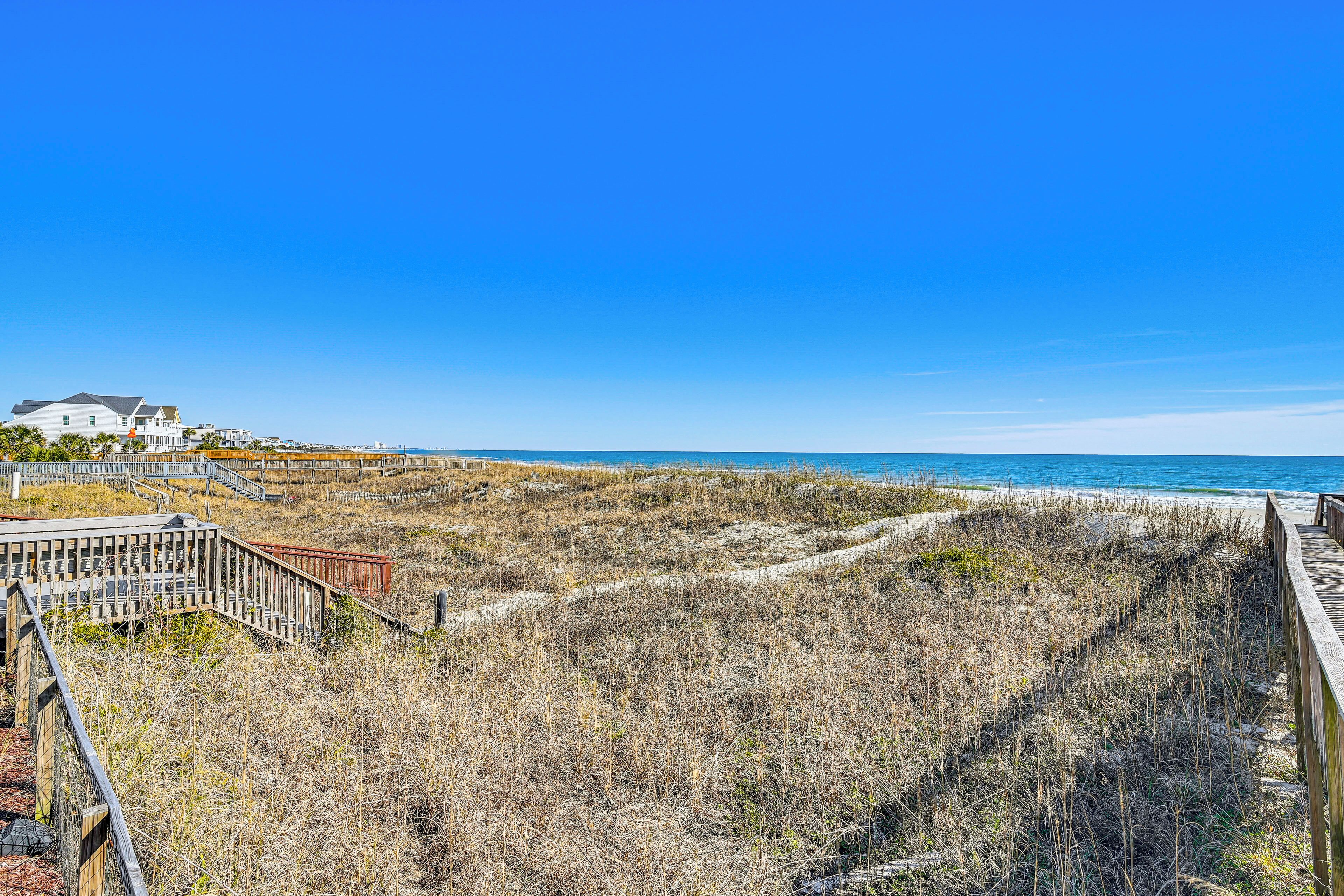 A beach view with a wooden boardwalk leading to a white building with a sloped roof.