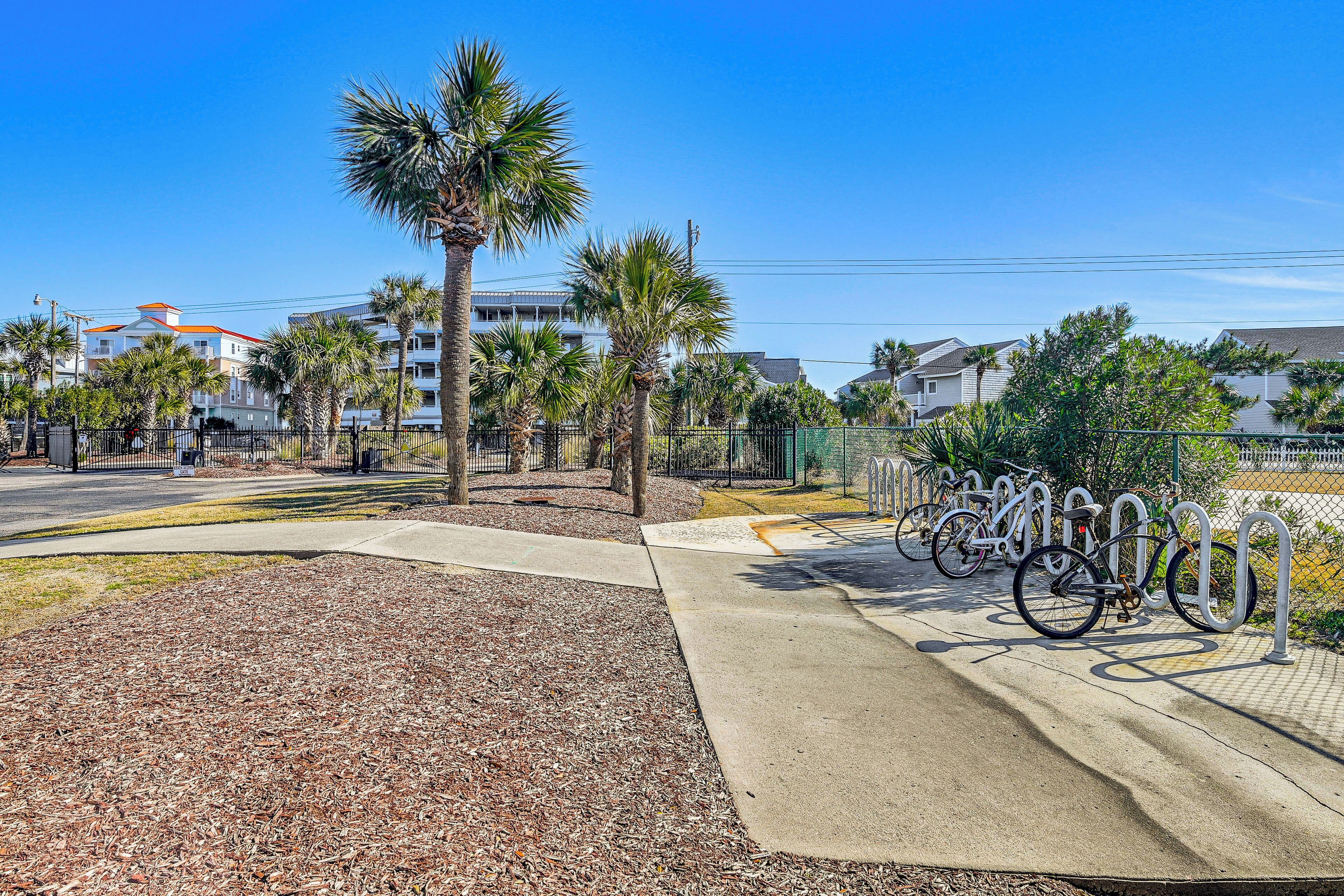 A paved bike parking area with metal bike racks, surrounded by palm trees and a sidewalk.