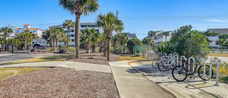 A paved bike parking area with metal bike racks, surrounded by palm trees and a sidewalk.