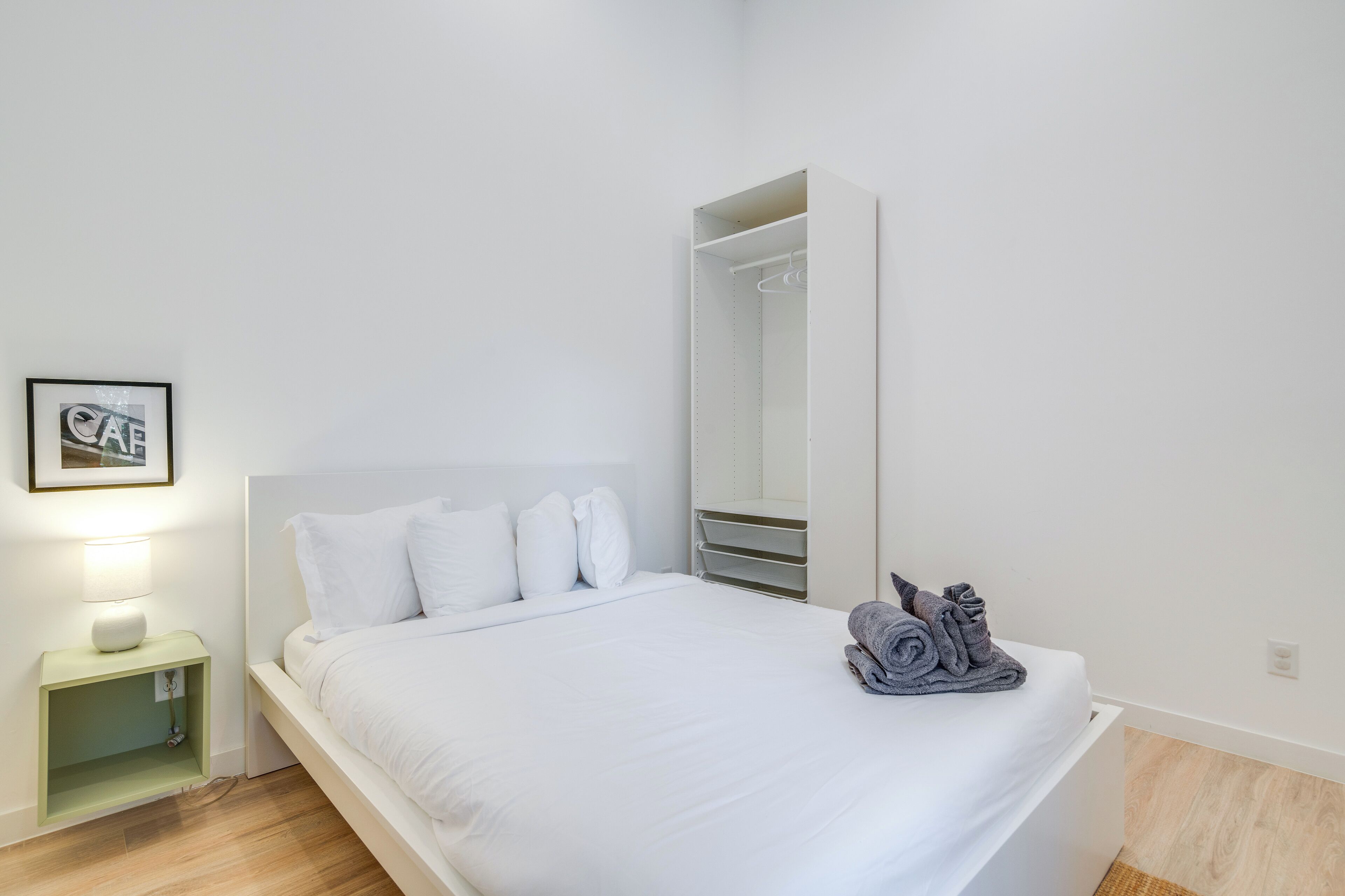 Una cama bien hecha con ropa de cama blanca, una lámpara de noche, una mesita de noche pequeña verde, un espejo y una toalla enrollada sobre la cama.
