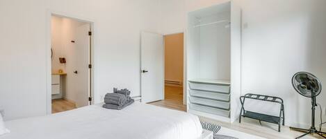 A modern bedroom with a large bed, a walk-in closet, a standing fan, and a small seating area.