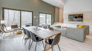 A modern living room with a large dining table, grey sofas, and a fireplace. - Hot Tub + Fire Pit: Modern North Carolina Retreat (Pisgah Forest)