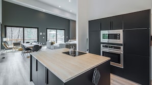 A modern kitchen with a central island, built-in appliances, and a dining area with a view of trees. - Hot Tub + Fire Pit: Modern North Carolina Retreat (Pisgah Forest)