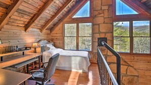 Un'accogliente camera da letto con soffitto in legno, pareti in pietra, un letto grande, una scrivania con una lampada e vista sulla foresta dalle finestre.