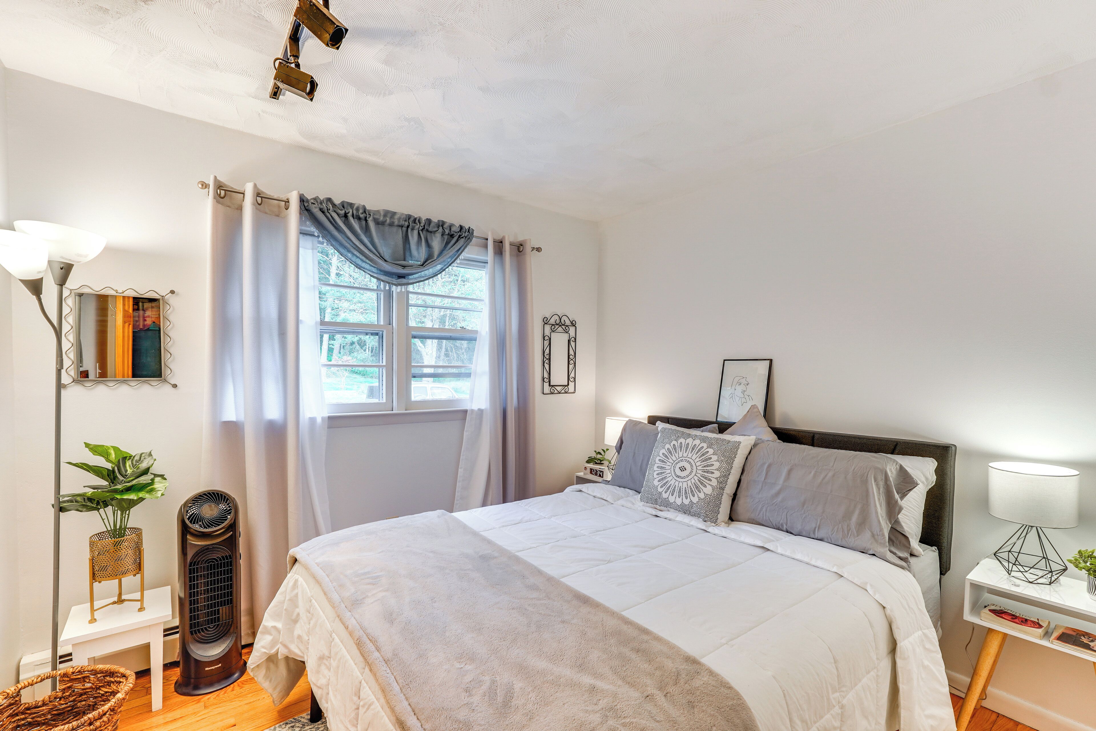 A bedroom with a bed, bedside tables, a standing fan, a window with curtains, and a wall-mounted lamp.