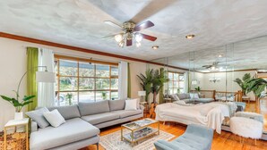A spacious living room with a grey sofa, a coffee table, and a blue armchair. There is a ceiling fan, a large window with curtains, and a mirror on the wall. - Creekside Catskills Retreat: Deck, Bbq, & Firepit (Parksville)