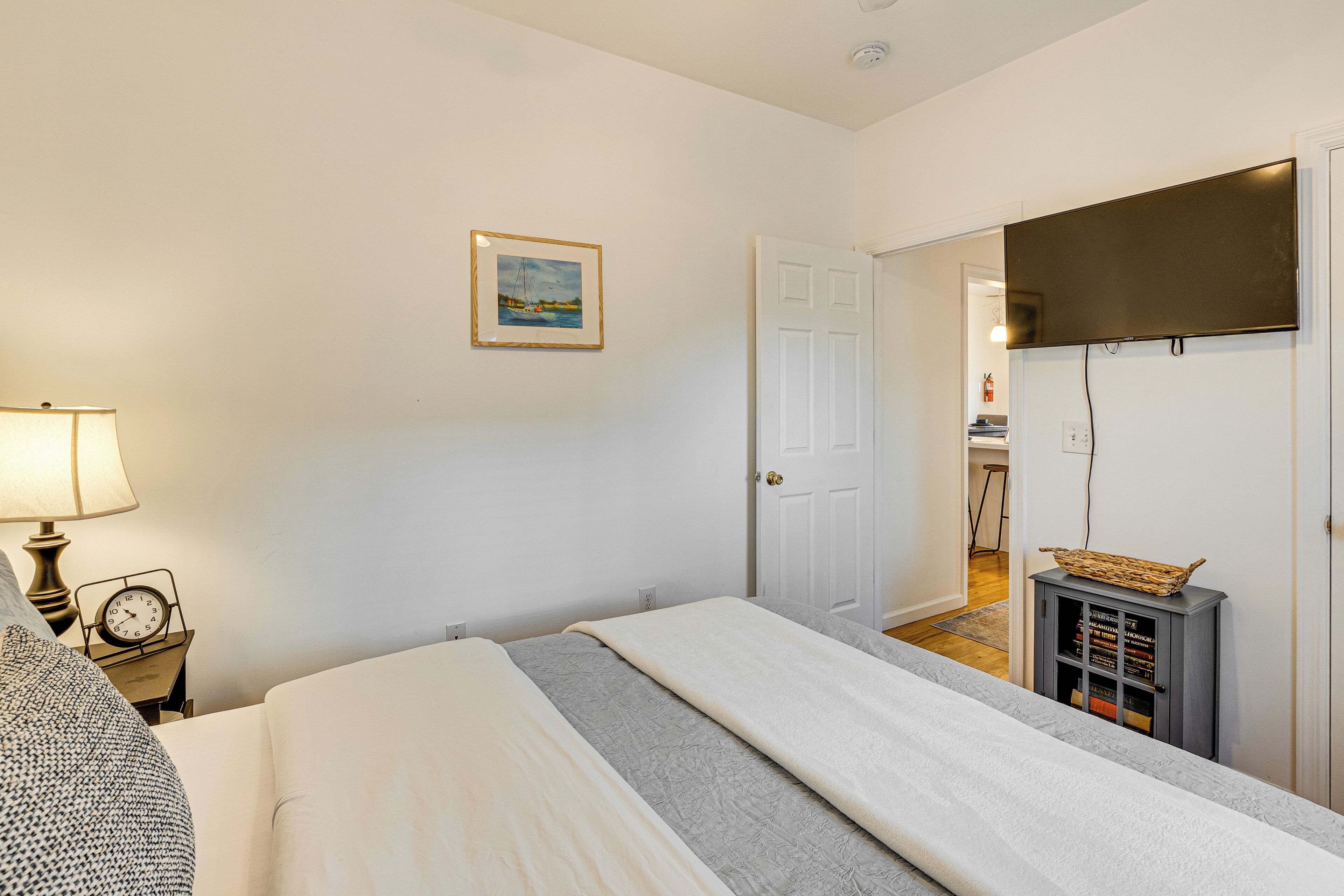 Une chambre à coucher avec un lit, une table de chevet avec une lampe et une horloge, un tableau encadré au mur et une porte donnant sur une autre pièce.
