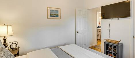 Une chambre à coucher avec un lit, une table de chevet avec une lampe et une horloge, un tableau encadré au mur et une porte donnant sur une autre pièce.