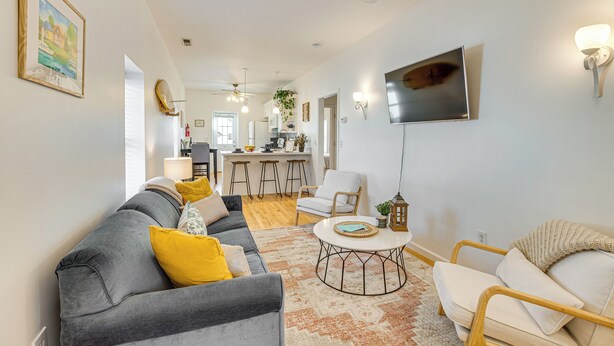 Una sala moderna con un sofá gris, un sillón color beige y una mesita de centro. Se observa una cocina al fondo con una barra y taburetes.