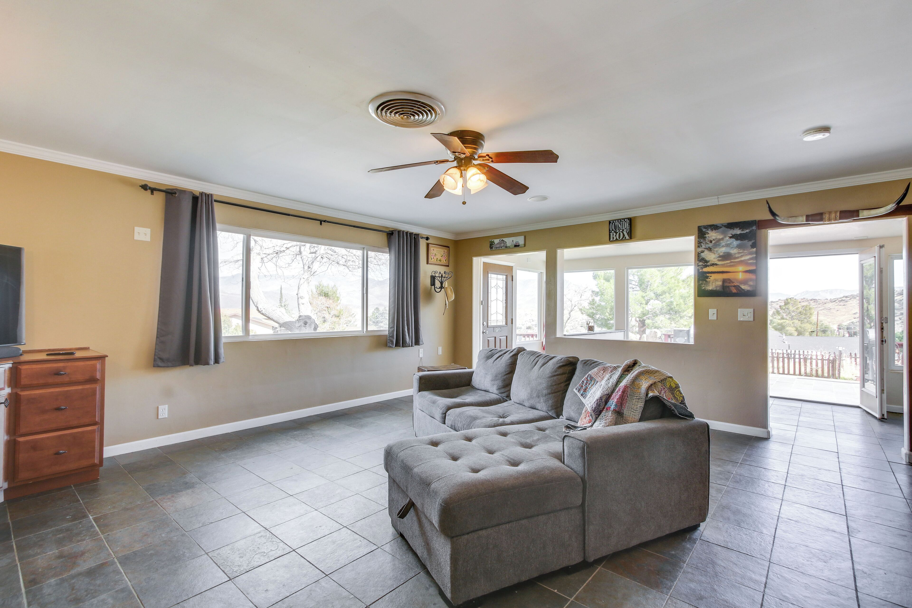 A spacious living area with a gray sectional sofa, a ceiling fan, and large windows offering a view of the outdoors.