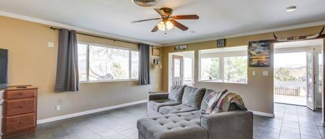 A spacious living area with a gray sectional sofa, a ceiling fan, and large windows offering a view of the outdoors.
