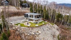 A modern two-story house with a glass facade, surrounded by a rocky landscape and trees. - Ski Chalet with Sauna | Mountain | L'Elegant (Saint-Côme)