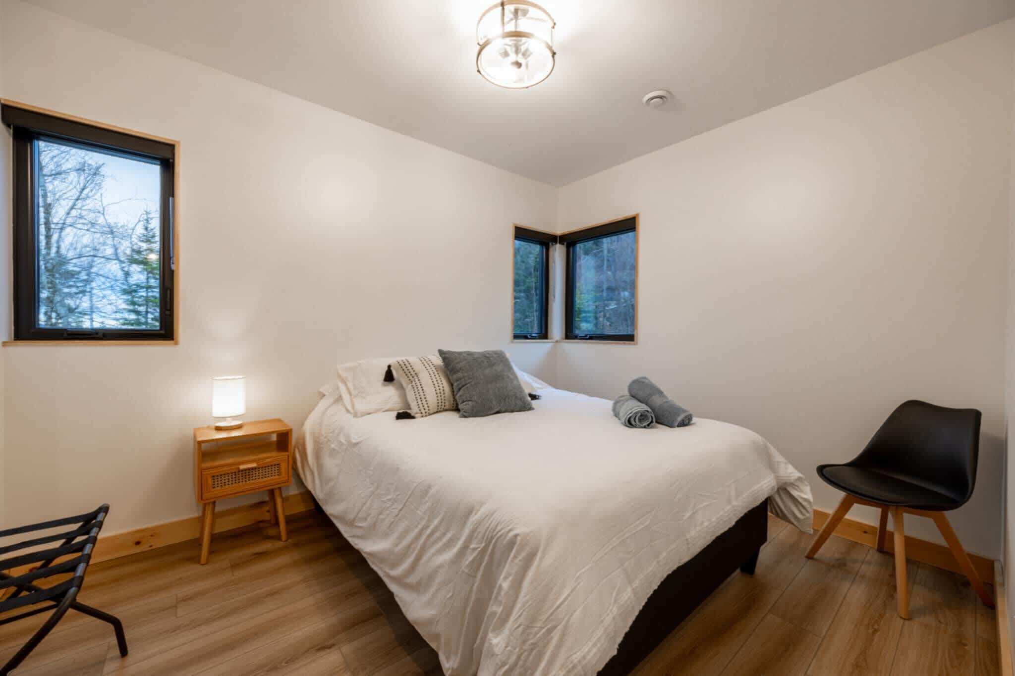 Une chambre à coucher avec un lit, deux fenêtres, une table de chevet avec une lampe, une chaise et un plancher en bois.