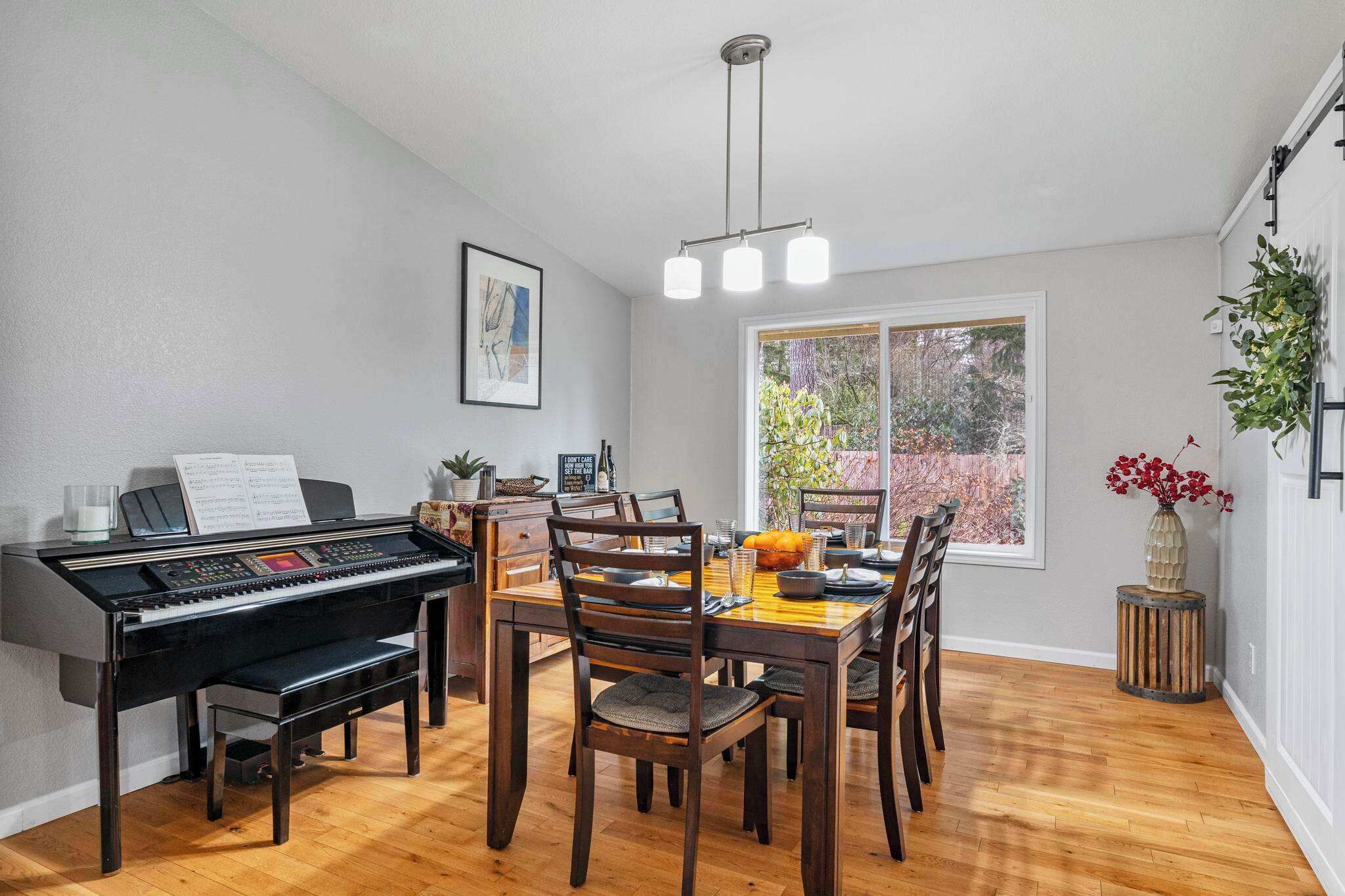 Uma sala de jantar com um piano, cadeiras de madeira, uma mesa com um arranjo de abóboras, uma janela com vista para árvores e um lustre.
