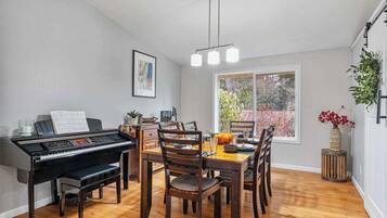 Uma sala de jantar com um piano, cadeiras de madeira, uma mesa com um arranjo de abóboras, uma janela com vista para árvores e um lustre.
