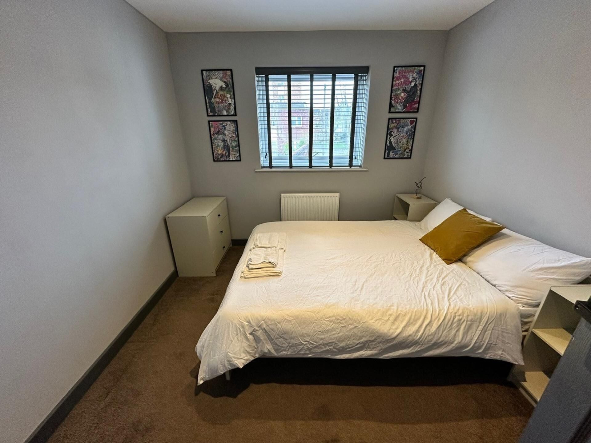 Un dormitorio con cama, un radiador, una ventana con persianas, una mesita de noche y un aparador.