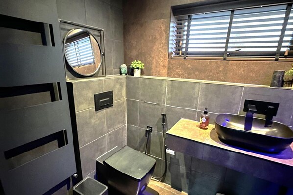 A compact bathroom with a modern design, featuring a round mirror, a small sink with a bowl, and a window with blinds.