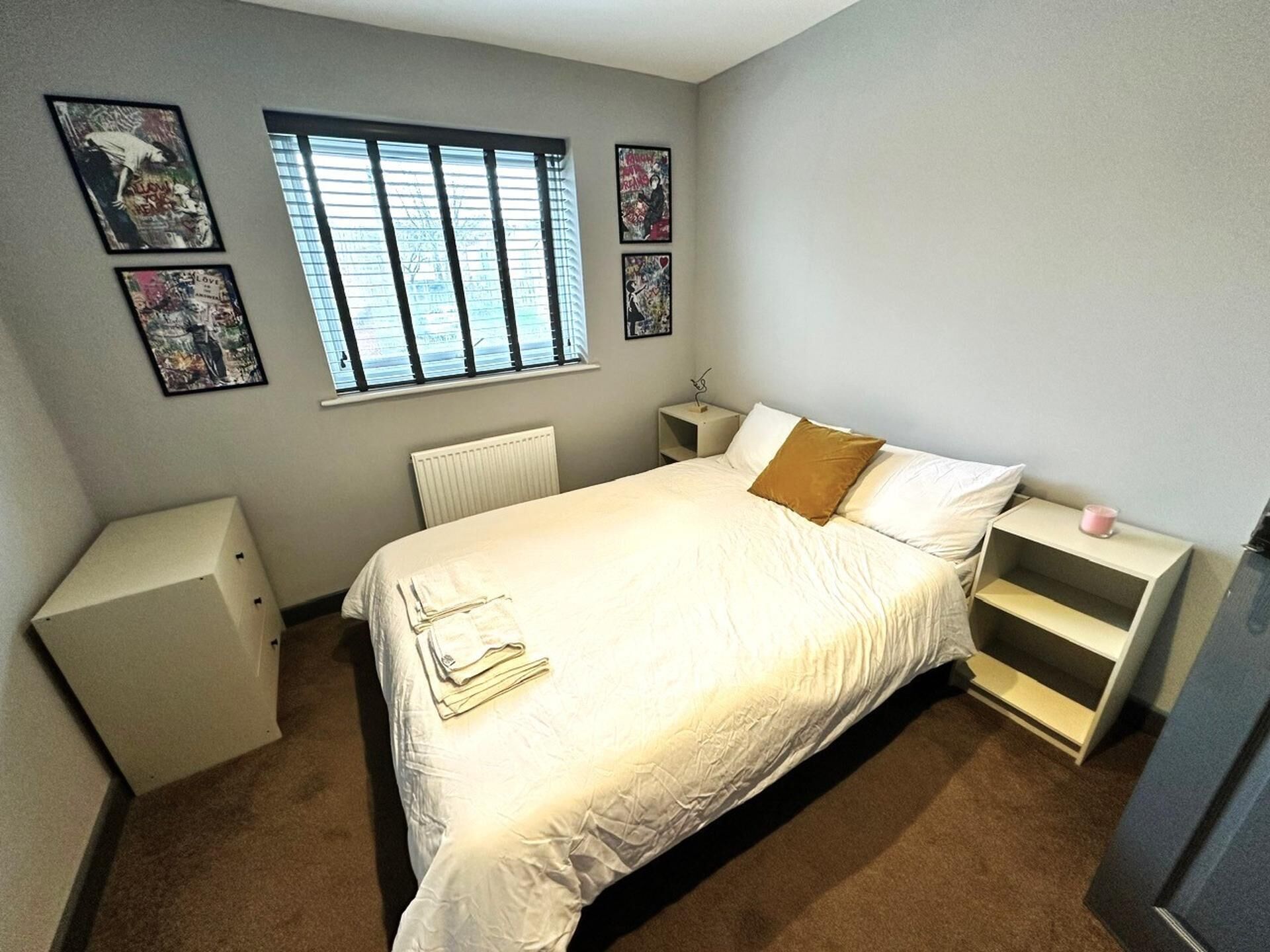A bedroom with a bed, a nightstand, a radiator, a window with blinds, and framed pictures on the wall.