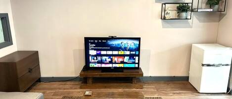 Una sala de estar con un televisor de pantalla plana sobre un soporte de madera, un refrigerador blanco y una pequeña estantería con plantas.