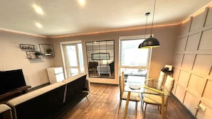 Ein modernes Wohnzimmer mit Holzboden, einer Couch, einem Fernseher, einem Essbereich mit einem Glastisch und Stühlen sowie einem großen Fenster.
