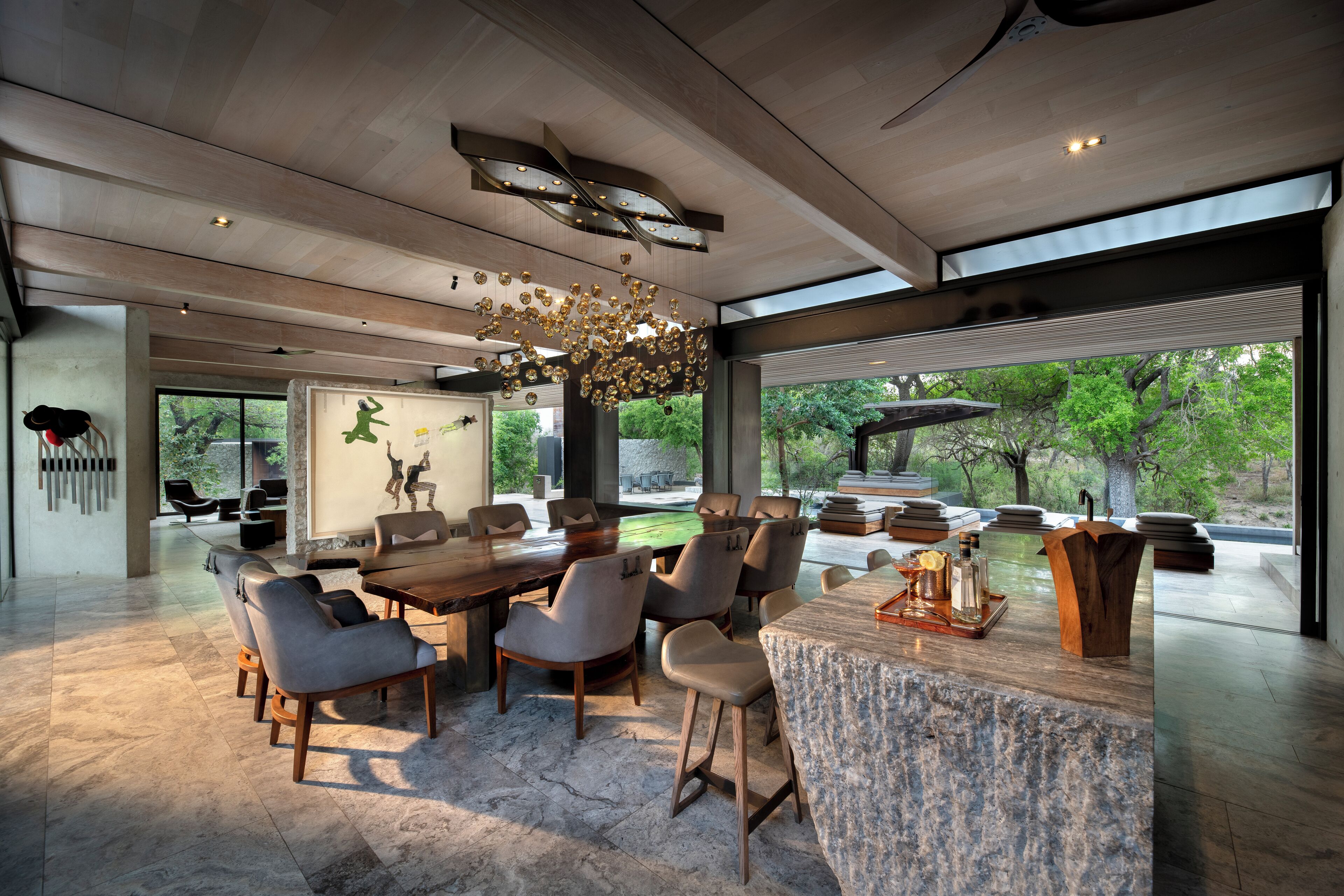A modern dining area with a large wooden table and comfortable seating, large windows offering views of greenery, and a unique ceiling light fixture.