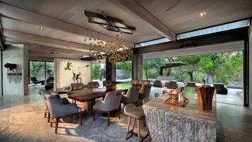 A modern dining area with a large wooden table and comfortable seating, large windows offering views of greenery, and a unique ceiling light fixture.