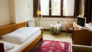 Une chambre d’hôtel avec un lit simple, un bureau avec un ordinateur, une chaise, une petite table avec une lampe et une fenêtre avec des rideaux.