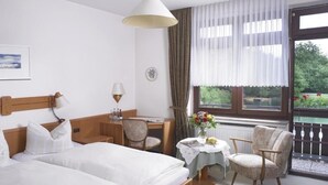Une chambre d’hôtel comprenant un lit, un bureau, deux chaises, une table avec un vase de fleurs et une fenêtre donnant sur un paysage verdoyant.