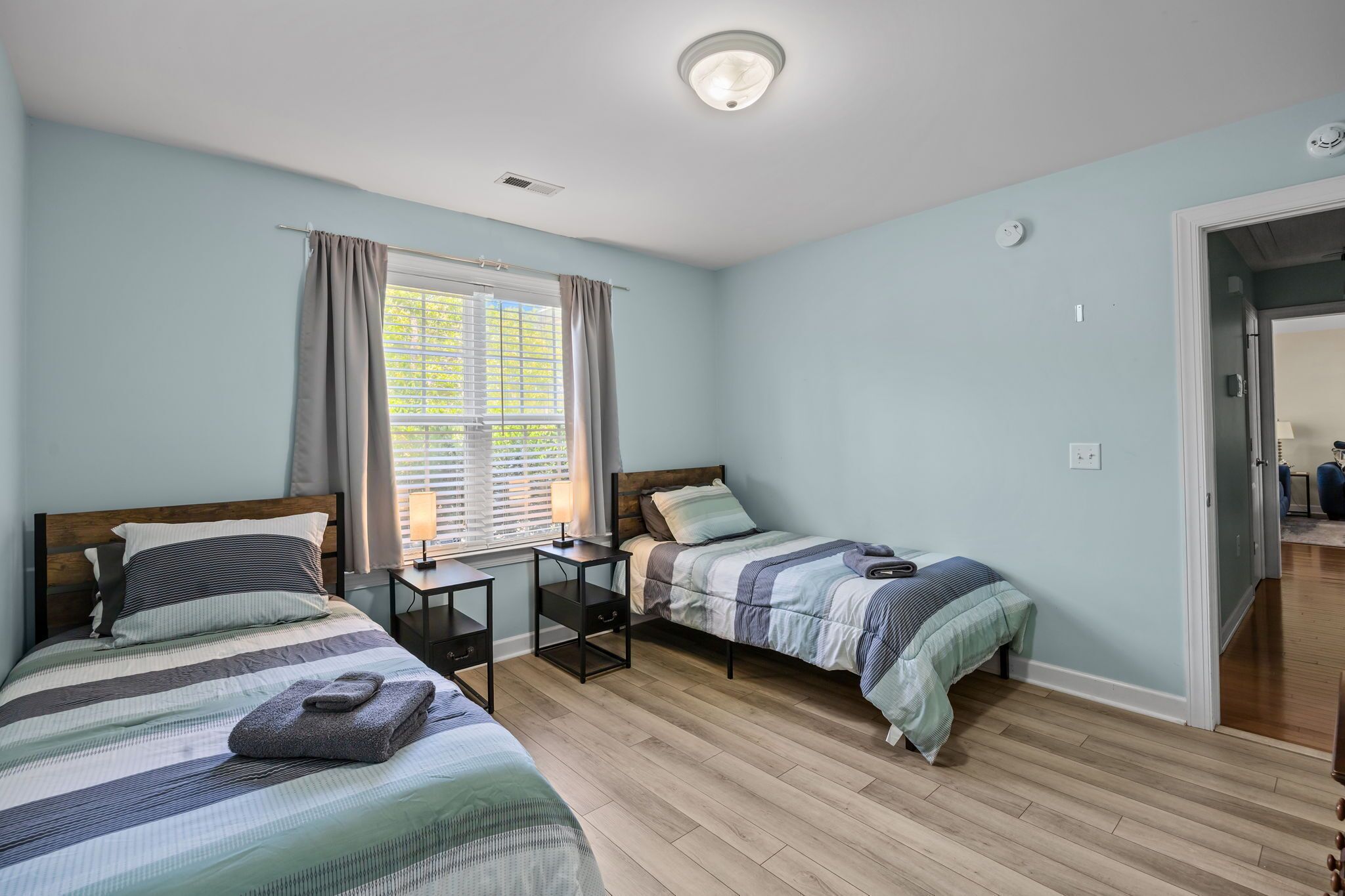 A bedroom with two beds, a window with curtains, a bedside table, and a lamp.