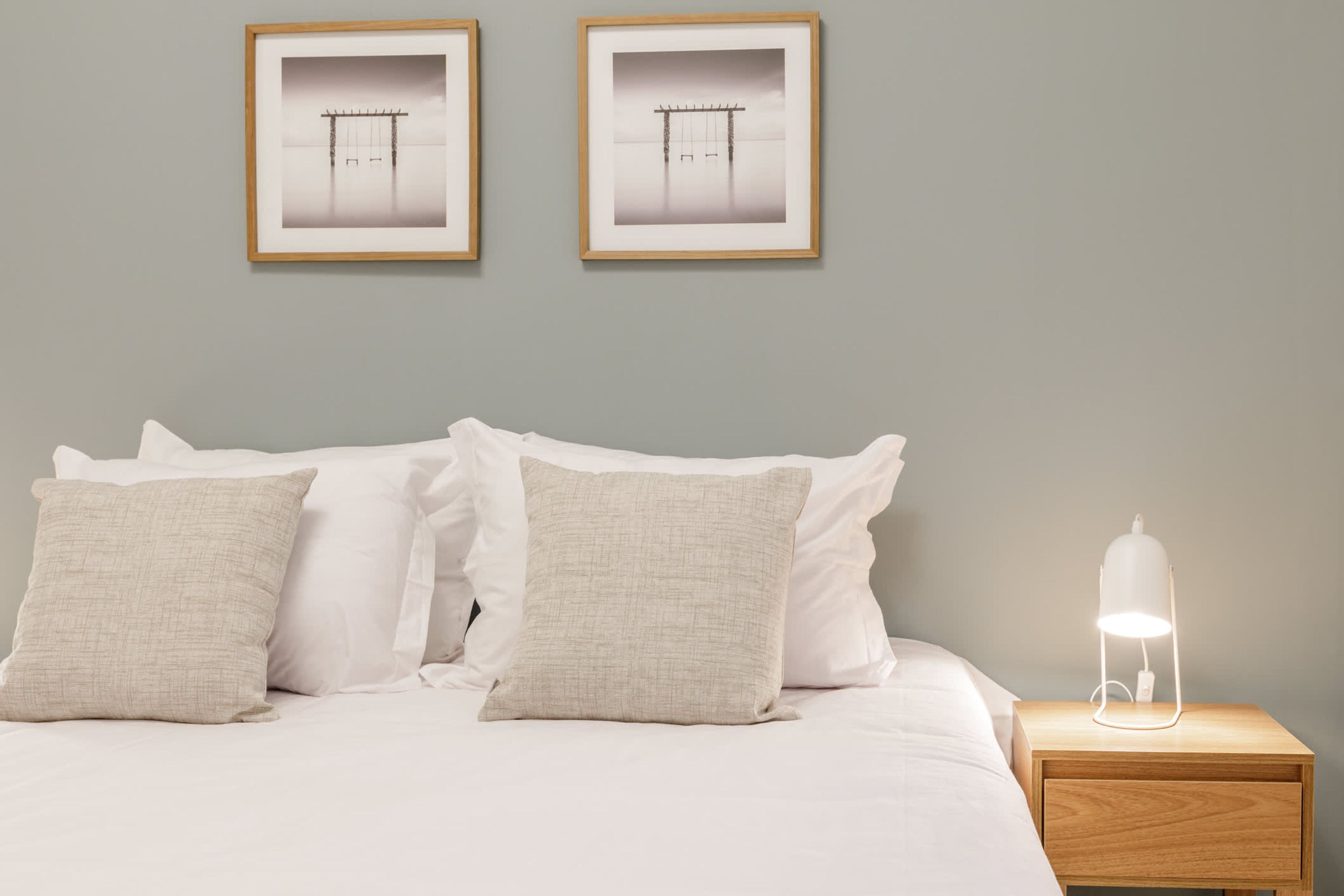 A neatly made bed with white bedding and two pillows, a bedside table with a lamp, and two framed pictures on the wall.