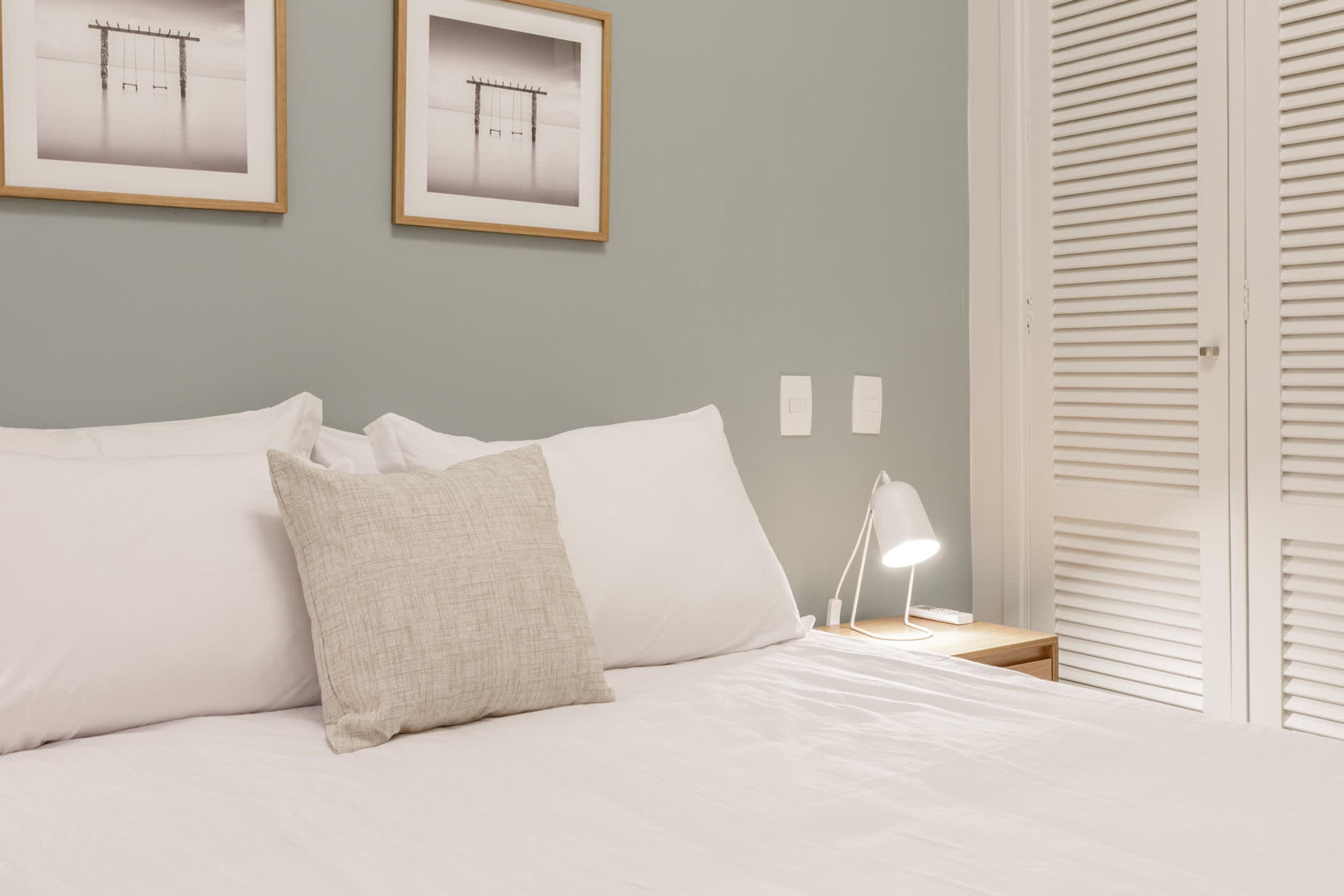 A neatly made bed with white linens and a beige pillow, a bedside table with a lamp, and a framed picture on the wall.