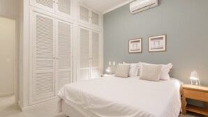 Une chambre à coucher avec un grand lit, du linge de lit blanc, une table de chevet avec une lampe et une armoire à portes à persiennes.