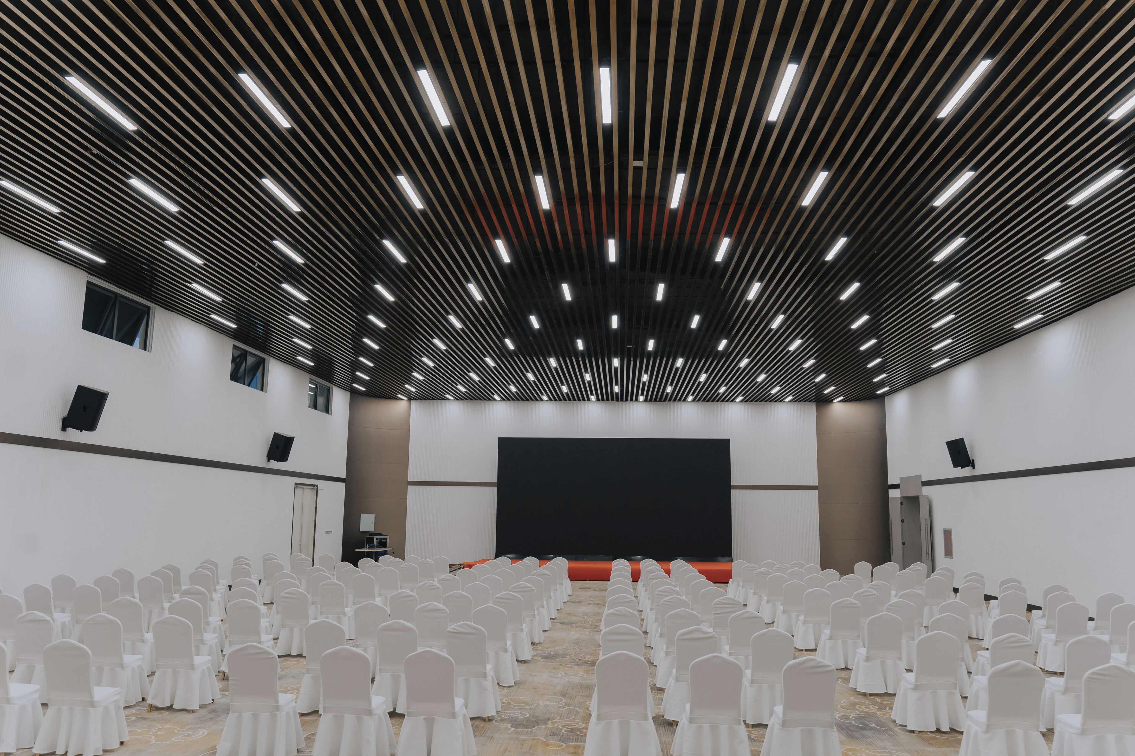 A conference room with rows of white chairs, a large black screen, and a modern ceiling with linear lighting.