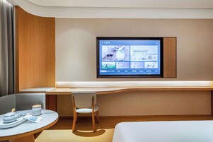 Una habitación de hotel moderna con un televisor de pantalla plana montado en la pared mostrando un menú, un escritorio de madera con una silla, una pequeña mesa redonda con un juego de té y una cama.