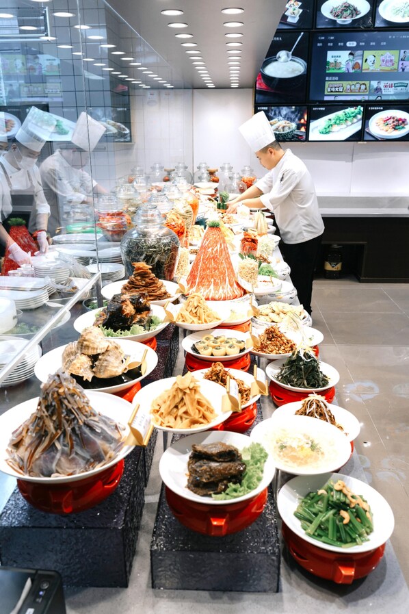 A buffet with various dishes arranged on a long table, a chef in uniform, and a digital menu display in the background. - Full Season Hotel (Qingdao)