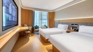 Una moderna camera d'albergo con una grande TV a parete, due letti, una scrivania e un angolo relax con vista sulla città.