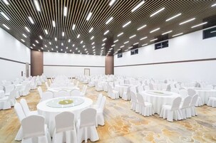 Un amplio salón de conferencias con mesas redondas y sillas blancas, techo alto con iluminación lineal y alfombra estampada.