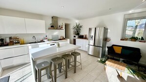 Une cuisine moderne dotée d’un îlot central, de tabourets de bar, d’un réfrigérateur et d’une fenêtre agrémentée de plantes.