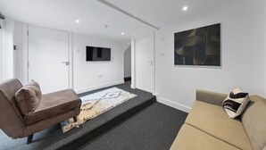 Un salón moderno con un sofá marrón, un sofá beige, un tapete estampado, un televisor de pantalla plana y una obra de arte geométrica en la pared.