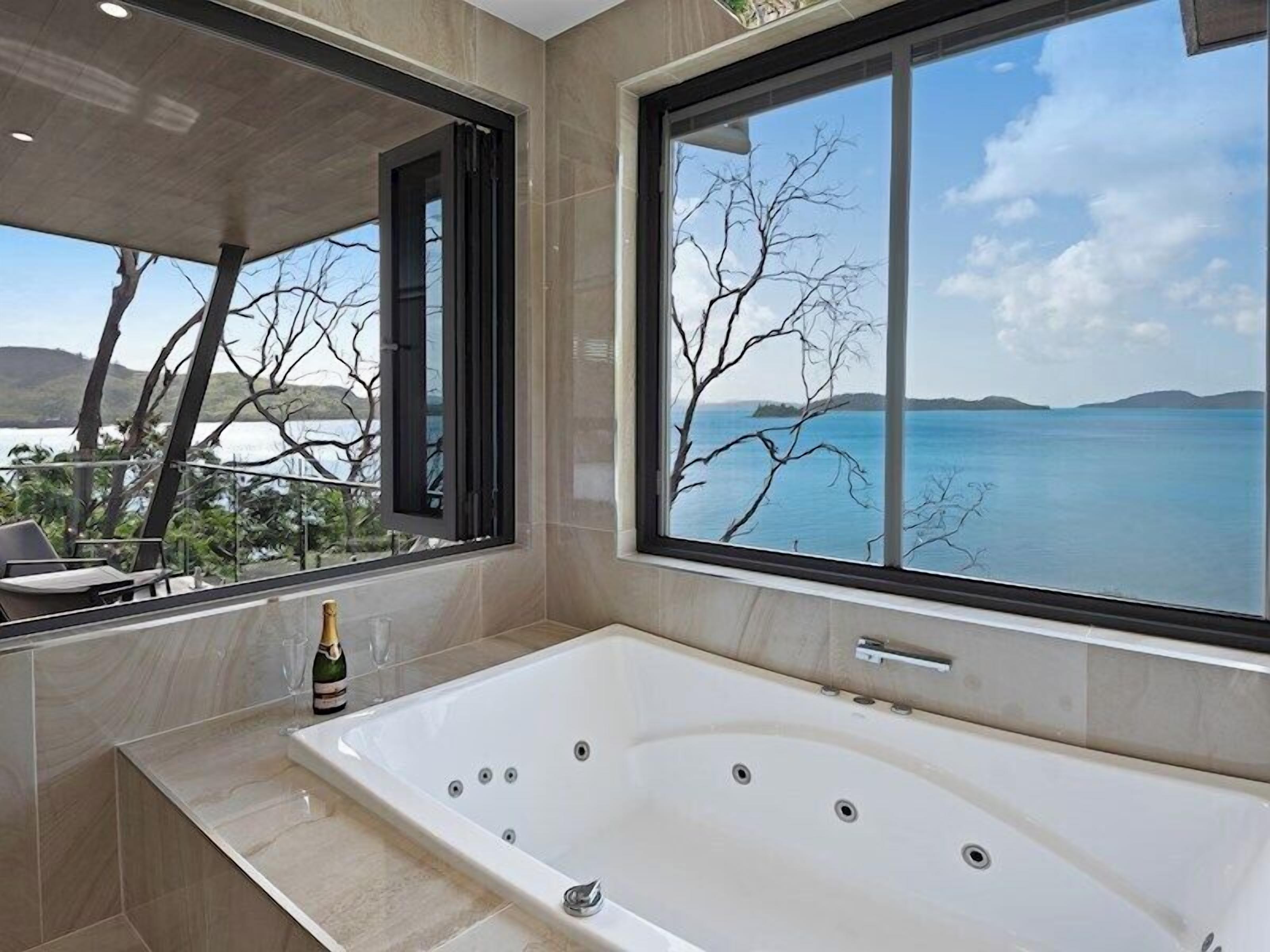 Une salle de bain moderne avec une grande baignoire, une bouteille de champagne et une vue sur un lac et des montagnes, visible à travers de grandes fenêtres.