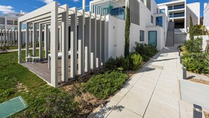 Un moderno edificio residencial con un diseño limpio y minimalista, destacando una fachada blanca prominente, un área de estar al aire libre cubierta y un jardín bien cuidado.
