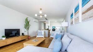 Una sala de estar moderna con un sofá, una mesa de centro y un televisor. Se observa una cocina al fondo.