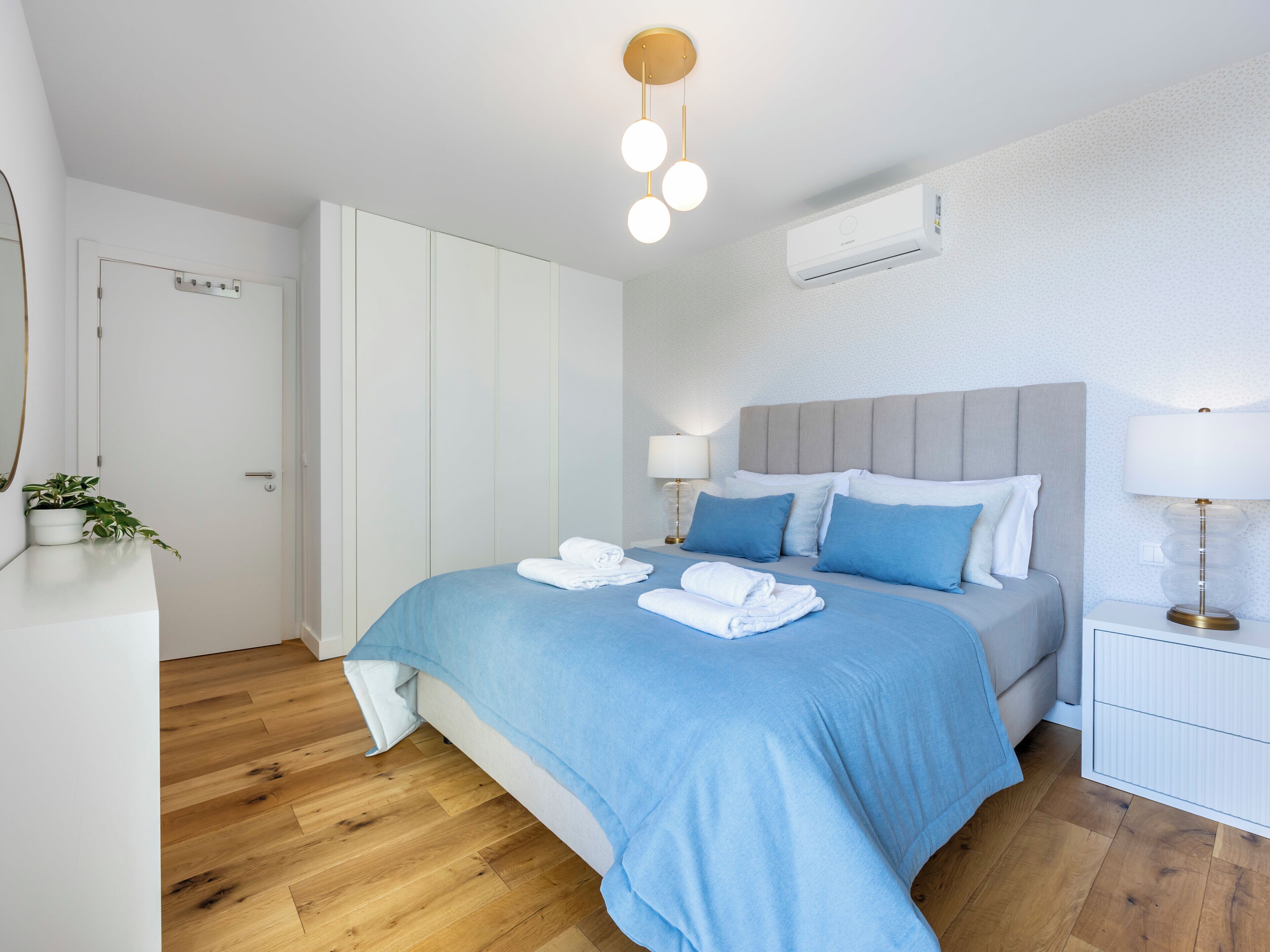 Et moderne hotelværelse med en pænt redt seng, et natbord med lampe, en vægmonteret aircondition og trægulv.