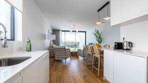 Una cocina moderna con armarios blancos, piso de madera, zona de comedor con sillas de mimbre y vistas amplias por las ventanas.