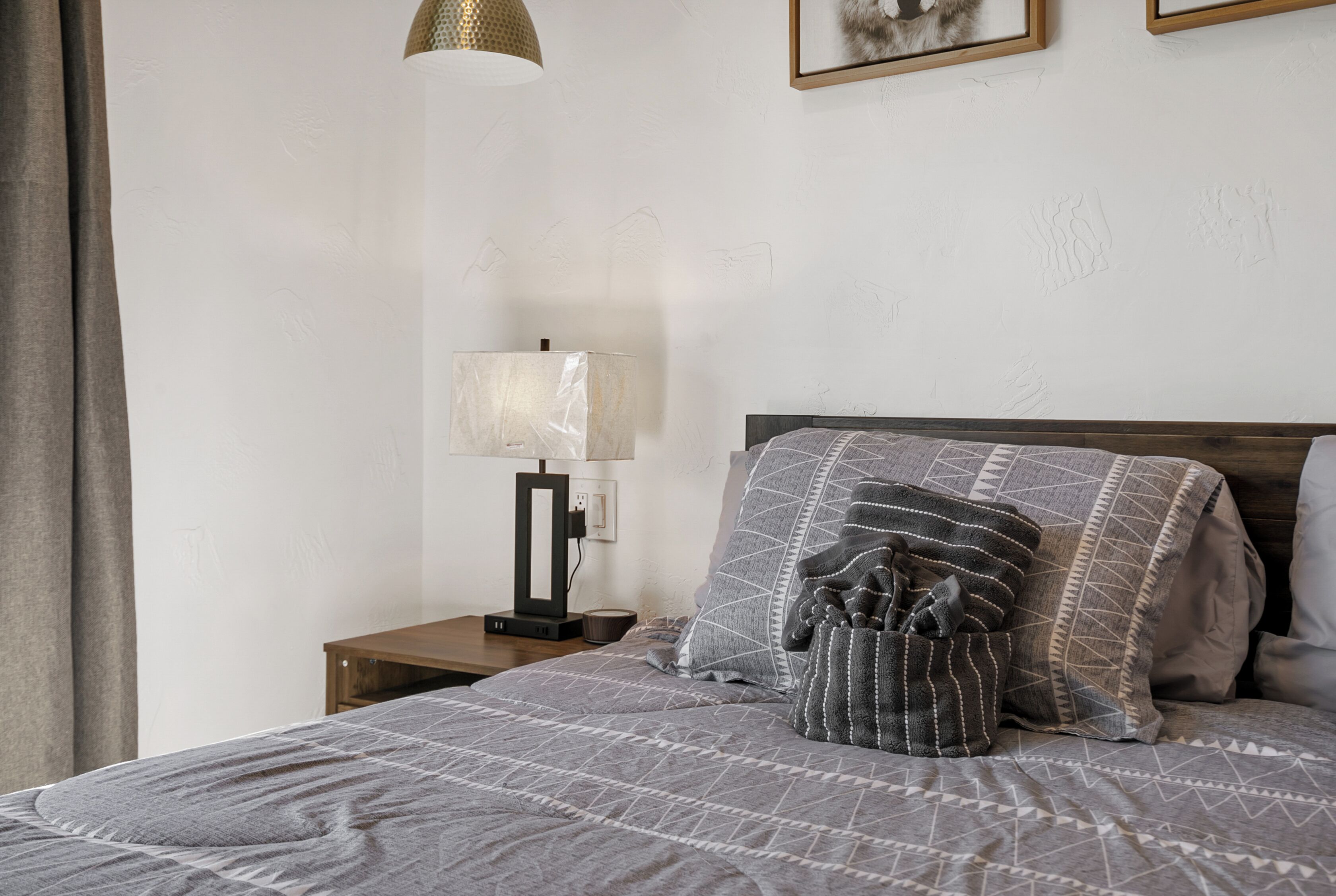 Ein ordentlich bezogenes Bett mit gemusterten Bettwäsche, ein Nachttisch mit Lampe und ein gerahmtes Bild an der Wand.