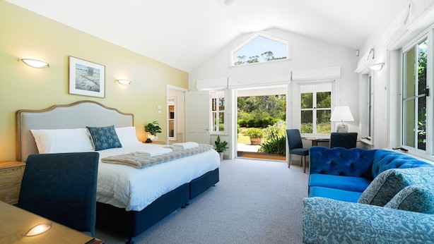 A bedroom with a large bed, a blue sofa, and a view of the garden through an open door. - Balmaghie - charming, secluded 7 acre rural retreat in the Southern Highlands (Bundanoon)